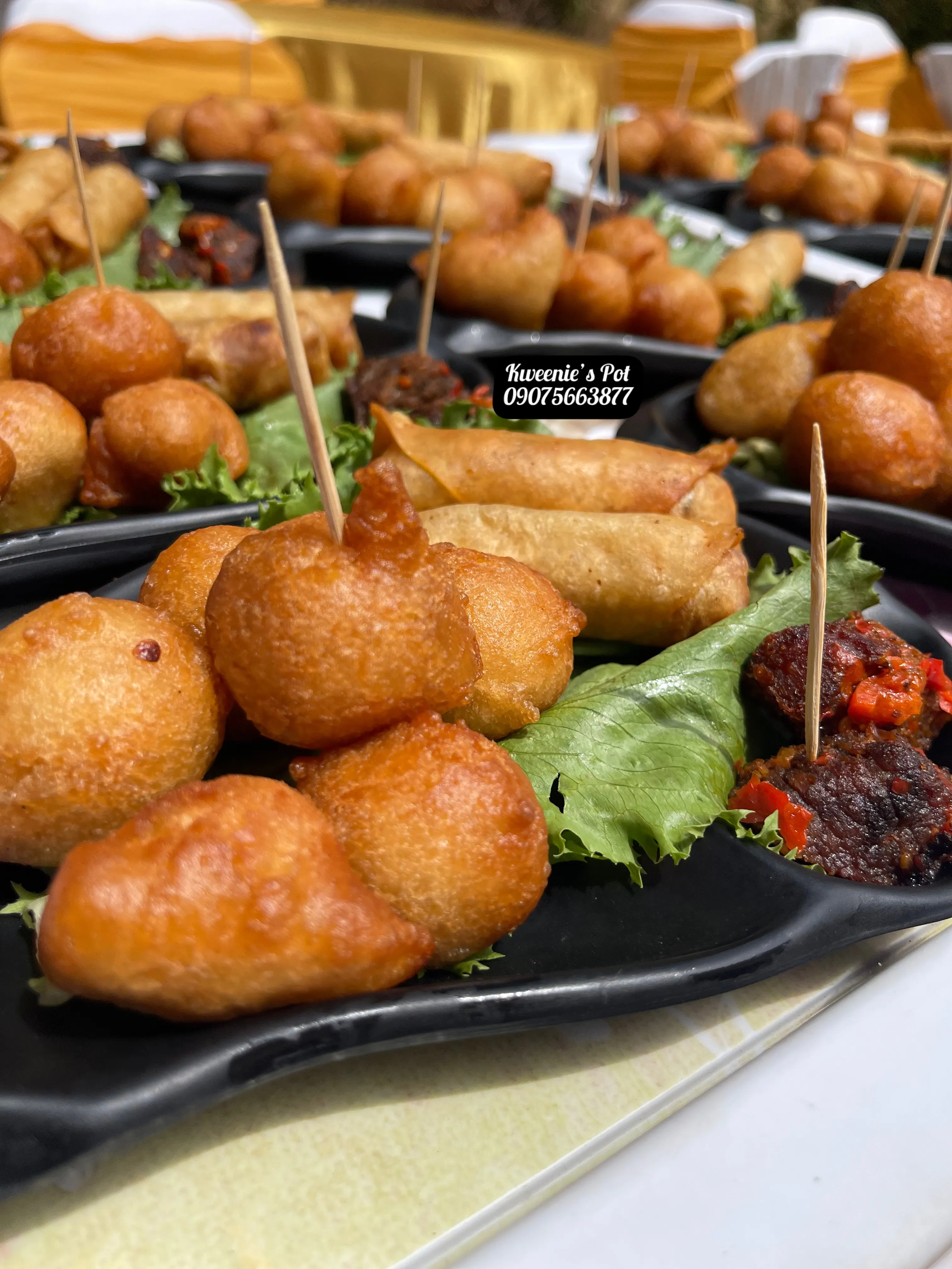 Assorted small chops with puff puff, spring rolls, and peppered meat on black plates at an event